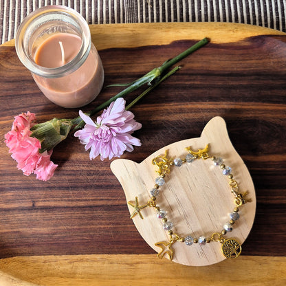 A beaded bracelet with gold-toned starfish charms and gray beads, displayed on a light wooden cat-shaped stand on a dark wooden tray, next to a pink flower and a glass jar candle.