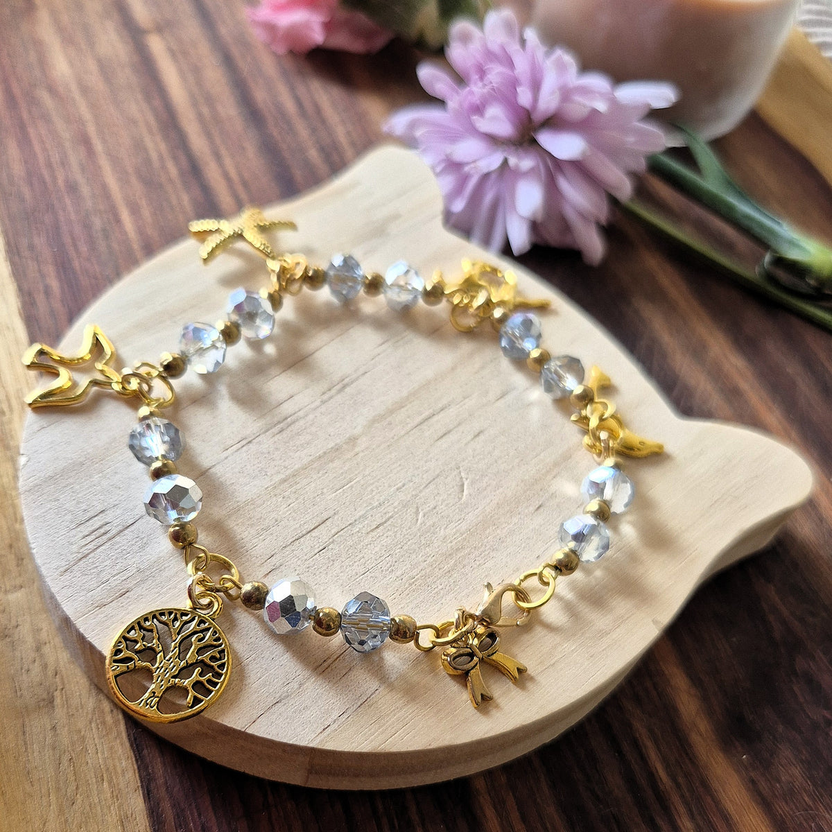 A gold-tone beaded bracelet with clear faceted beads, small gold charms, and a tree of life pendant, displayed on a wooden cat-shaped coaster.