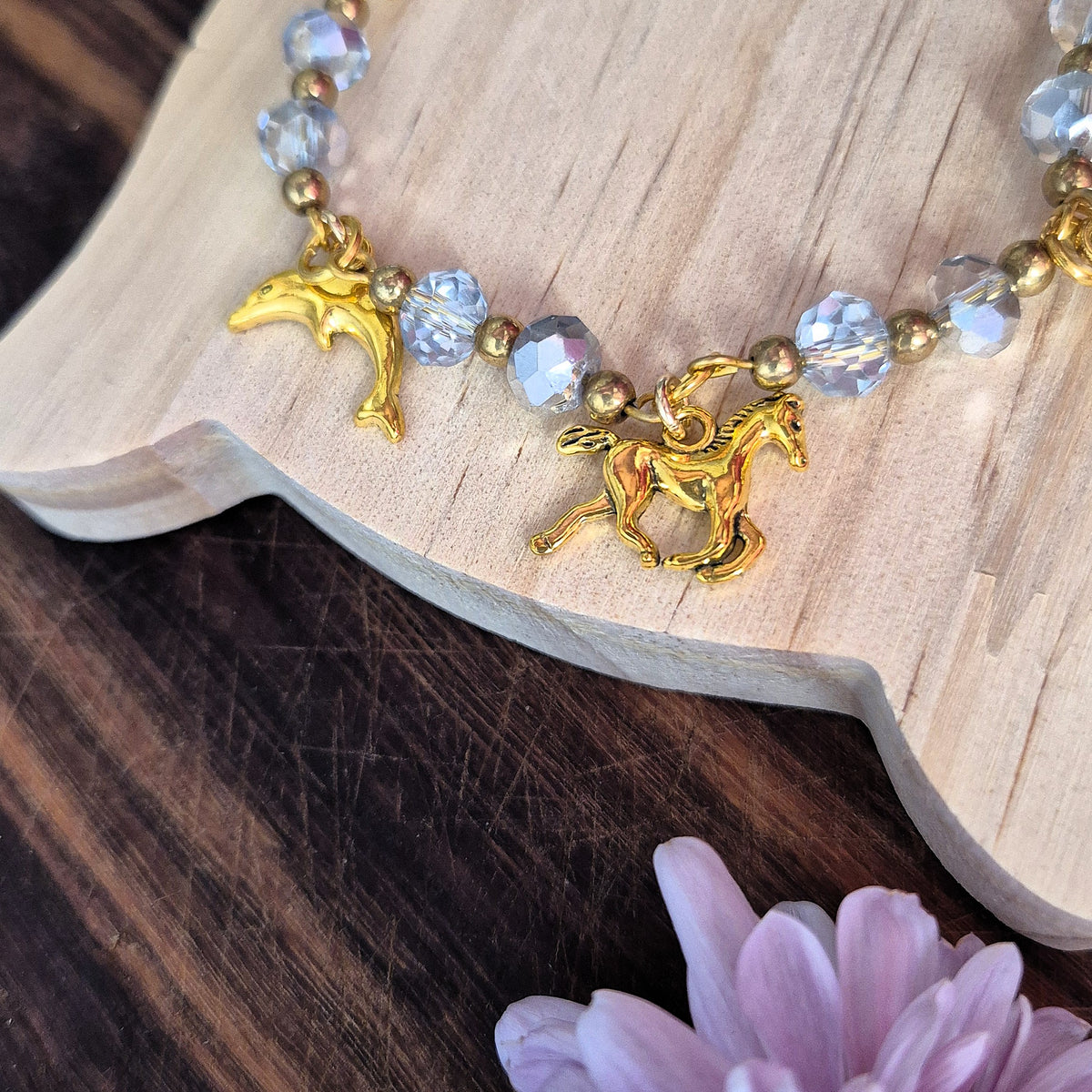 A close-up of a beaded bracelet with gold-toned unicorn and dolphin charms, clear faceted beads, and small metallic spacer beads, displayed on a light wood slice over a dark wood surface with a pale purple flower in the foreground.